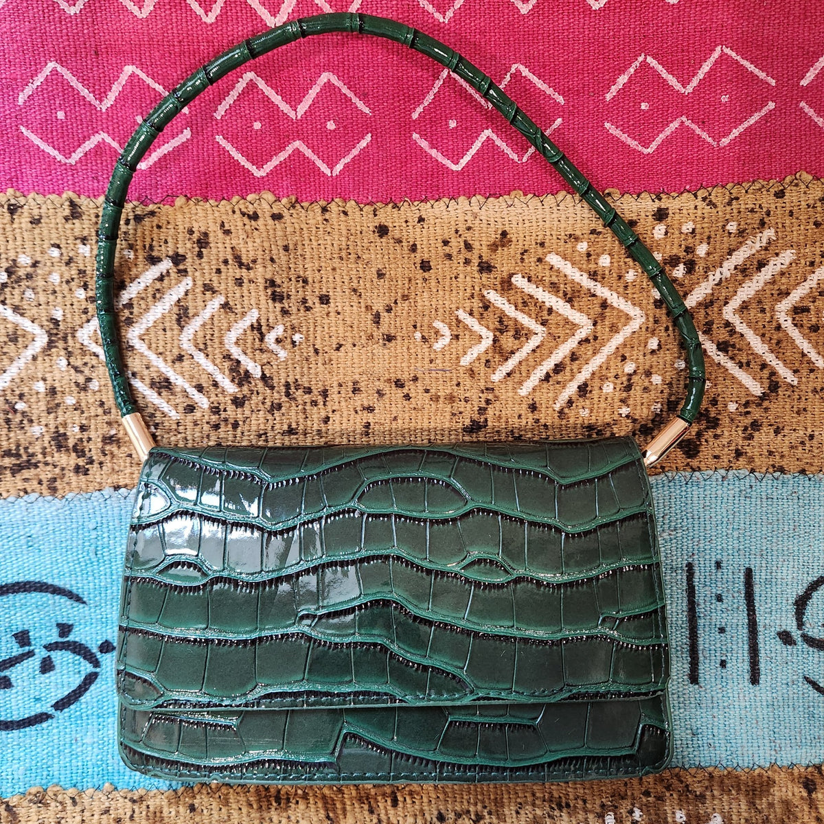 Forest green croc-embossed shoulder bag, front view on a patterned textile with shoulder strap arched above