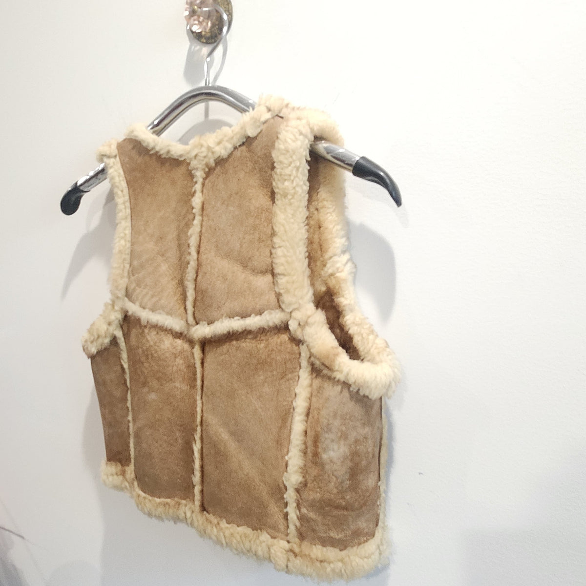 Lucille Golden Vintage, LLC tan suede shearling vest with cream wool trim hanging on a hanger against a white wall.