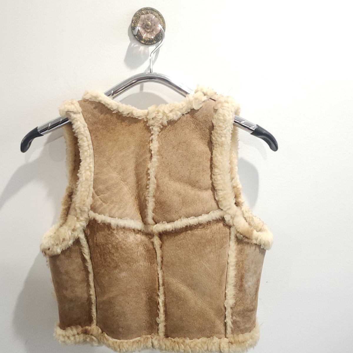 Lucille Golden Vintage, LLC tan suede shearling vest with cream wool trim hanging on a hanger against a white wall.