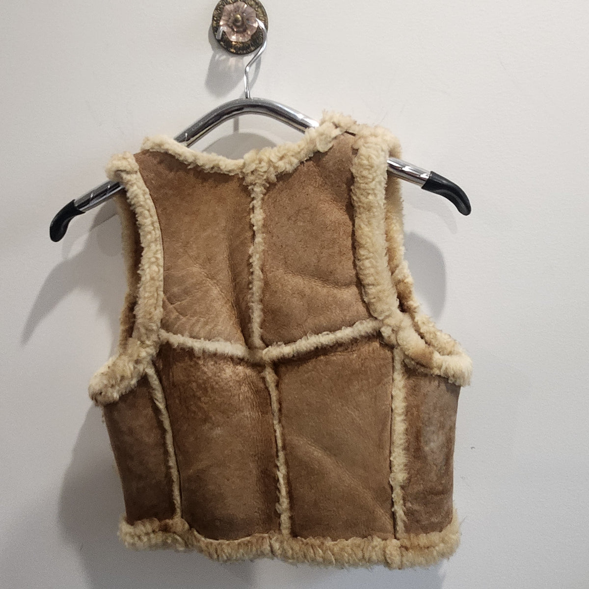 Lucille Golden Vintage, LLC tan suede shearling vest with cream wool trim hanging on a hanger against a white wall.