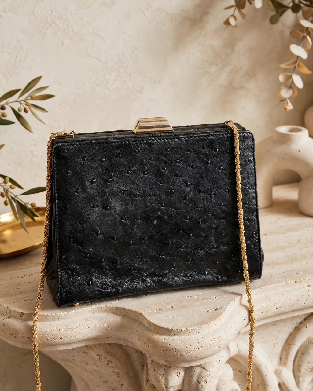 Editorial photograph of vintage Beoni Lucia black ostrich-textured leather evening bag with braided gold chain, styled on a sculptural stone table against a warm ivory backdrop